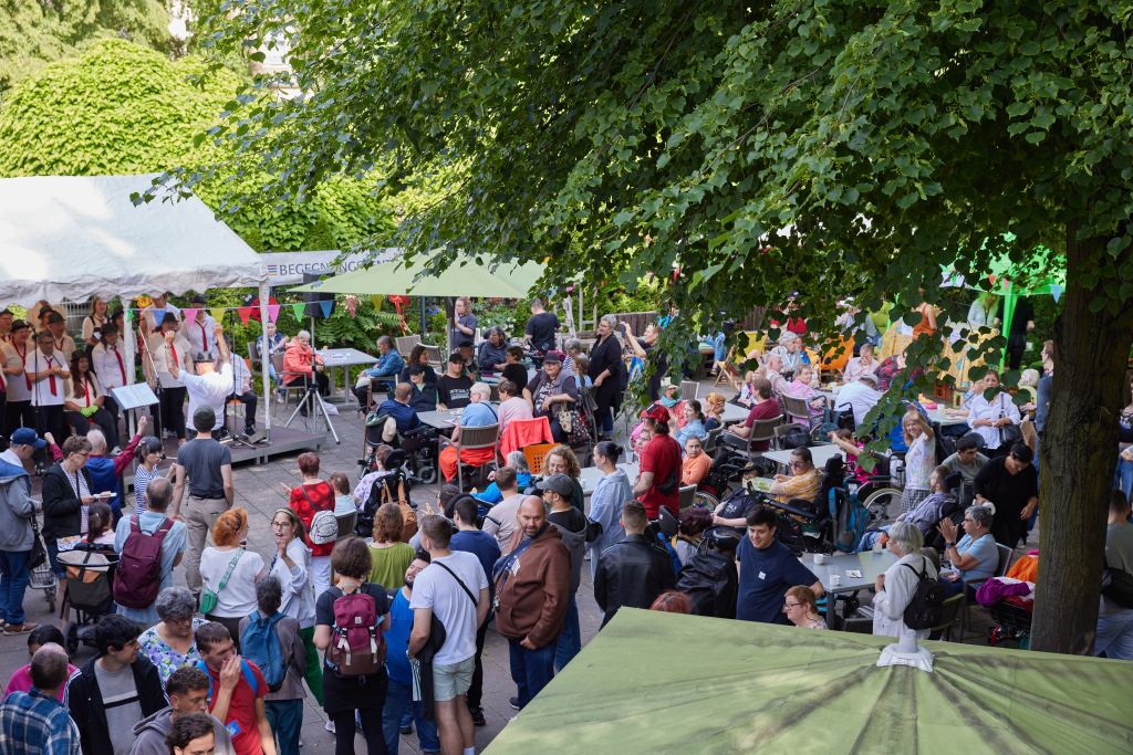 zahlreiche Menschen stehen uns sitzen vor einer aufgebauten Bühne, auf der ein Chor singt